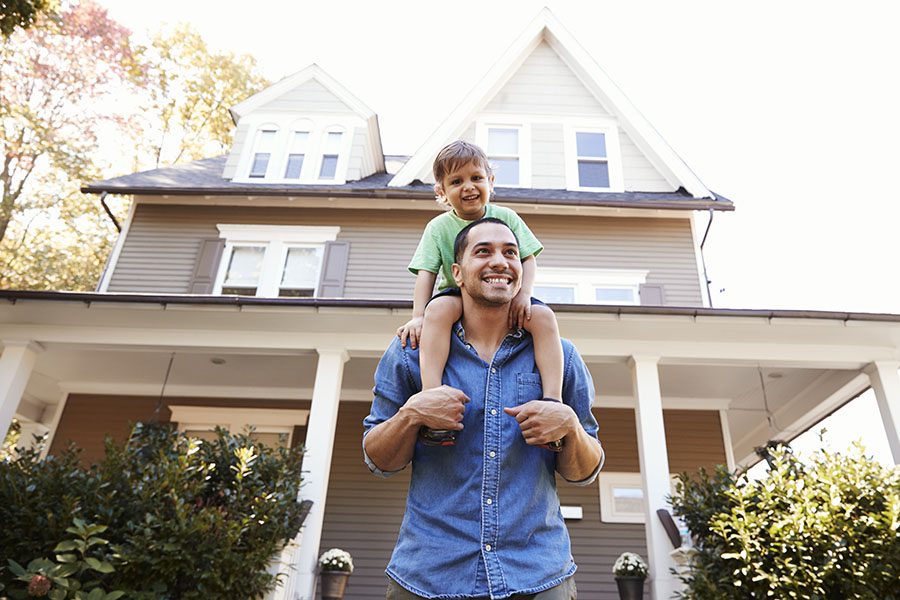 Personal Insurance - Portrait of a Cheerful Young Father Giving His Son a Piggyback Ride While Standing Outside Their Home